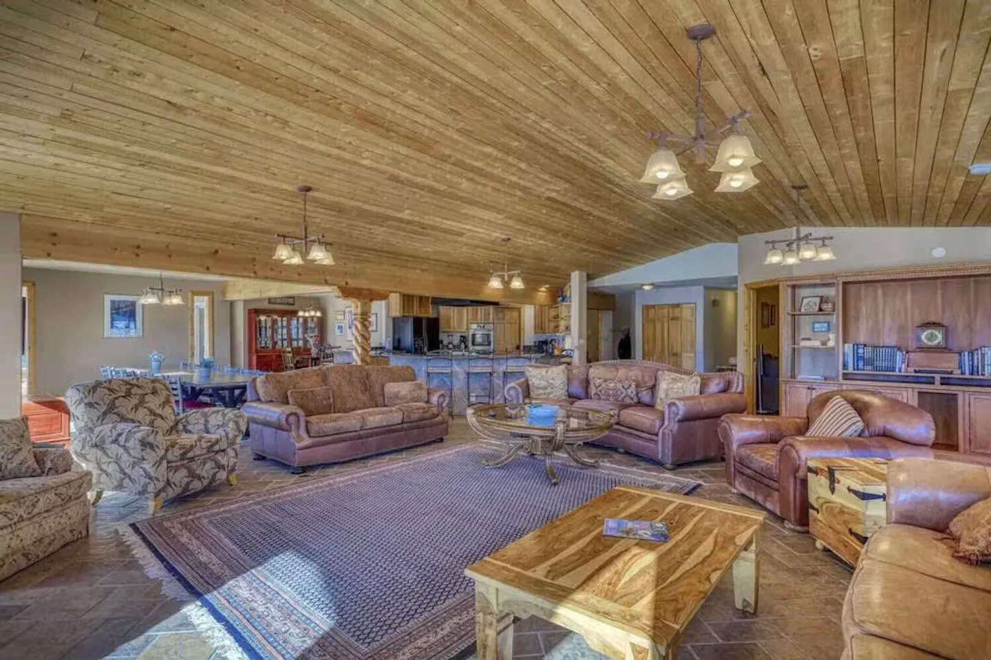 Open-concept living room with a wood-plank sloped ceiling, leather and fabric seating around coffee tables, and a view into the kitchen and dining areas.