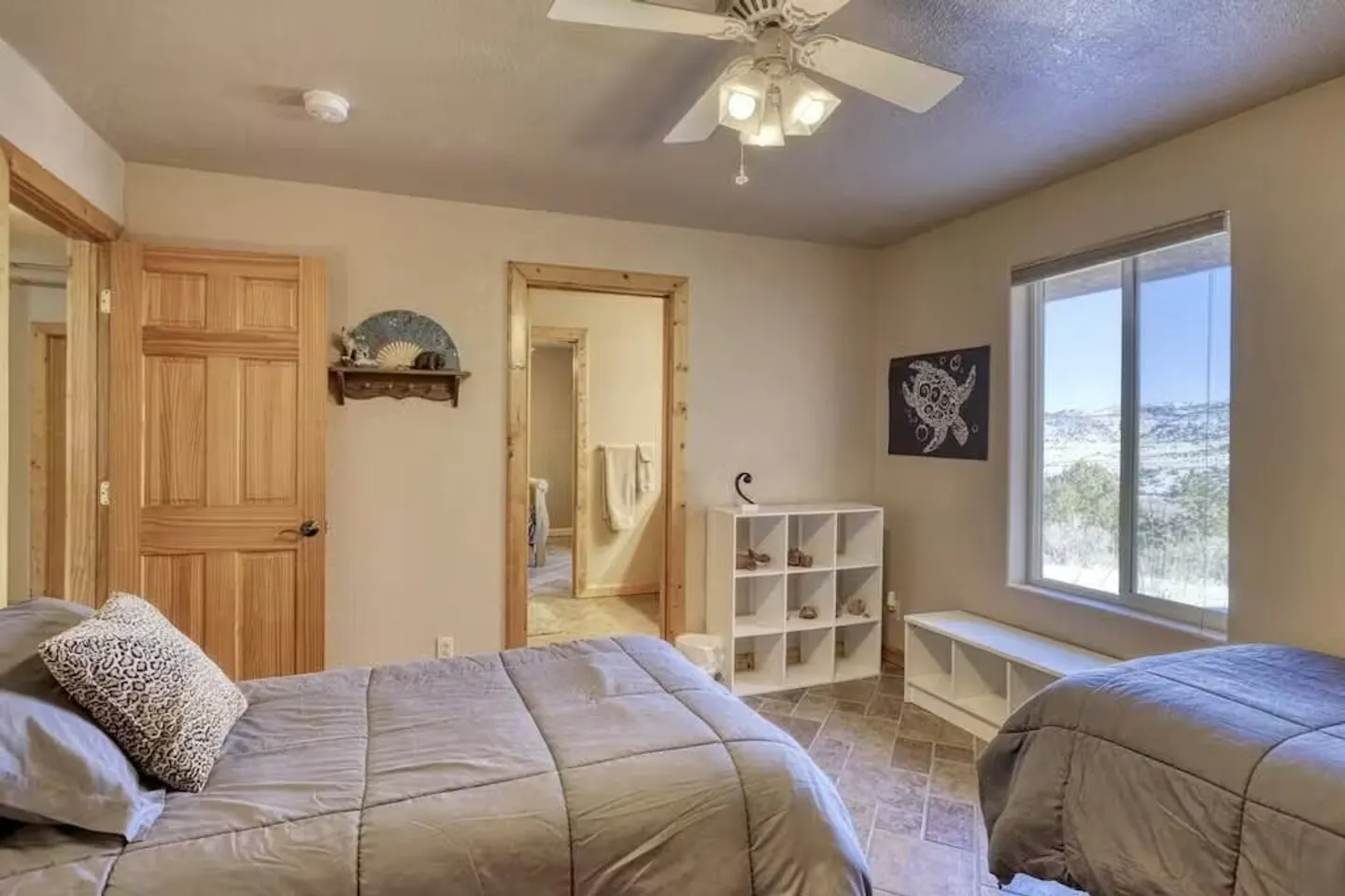Bedroom with two beds made up in grey bedding, tile flooring, white cubby shelving, a ceiling fan, and a window showing hills outside.