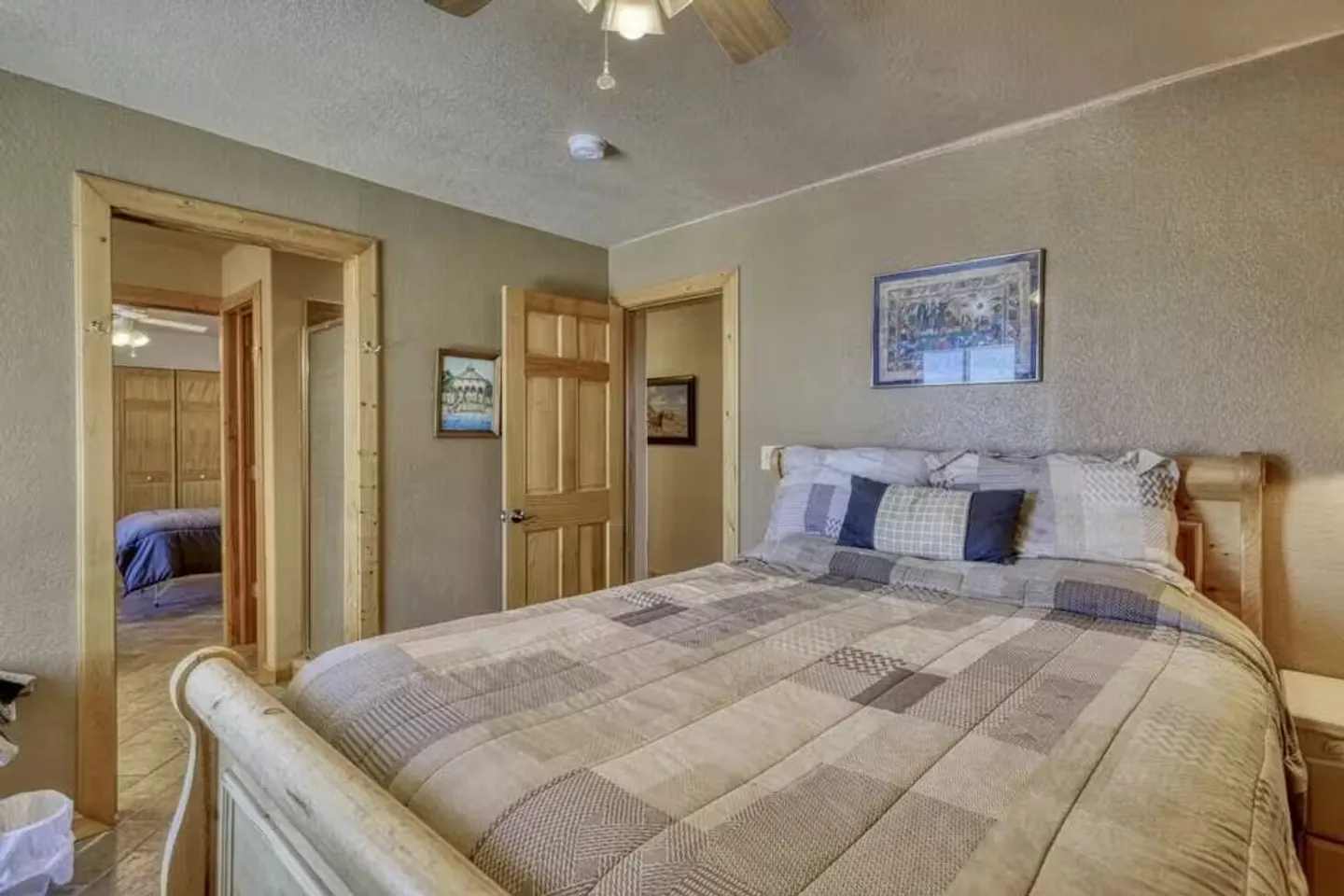 Bedroom with a wooden bed, patterned quilt, textured walls, ceiling fan, and open doorways showing adjoining rooms.