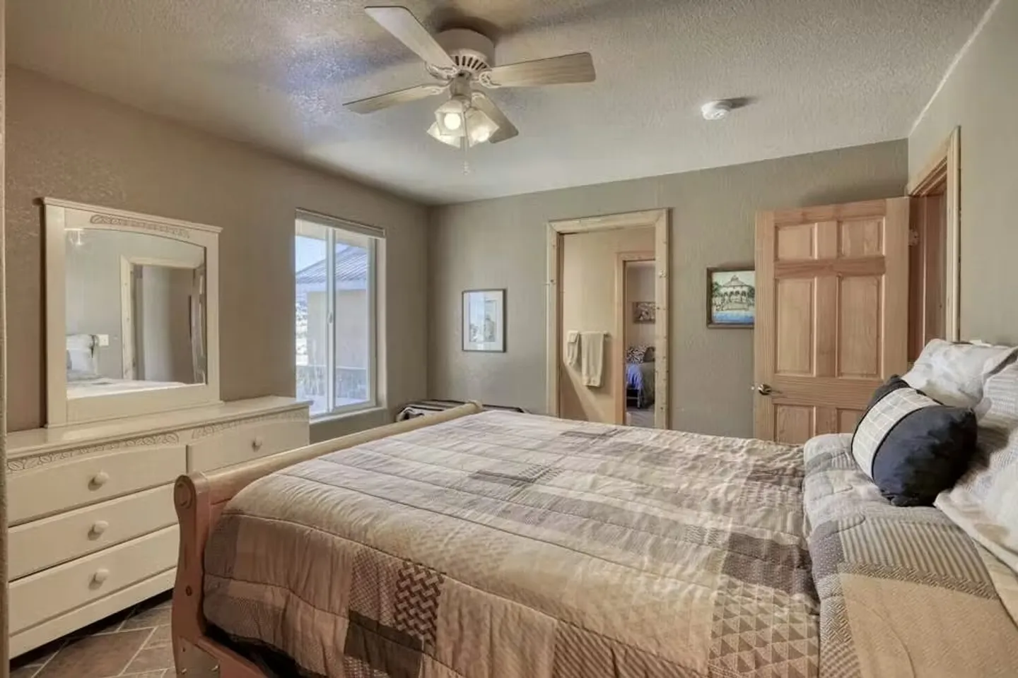Bedroom with a wooden sleigh-style bed, patchwork quilt, white dresser with mirror, ceiling fan, and doorways leading to adjoining rooms.