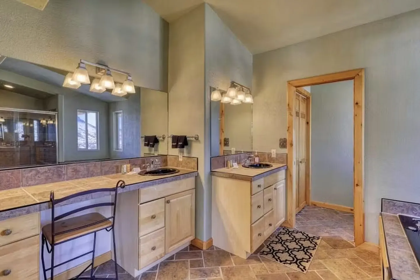 Primary bathroom featuring tiled countertops, two separate vanity areas with light wood cabinetry, tiled floors, and a view into an adjacent room.