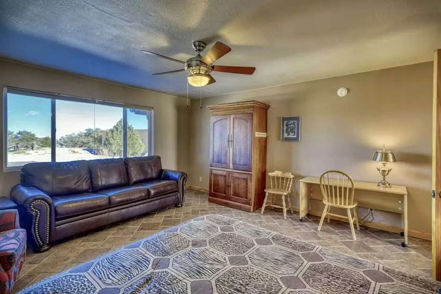 Bedroom or living area with a brown leather sofa, large window, wooden armoire, desk with lamp and chair, and a tiled floor with an area rug.
