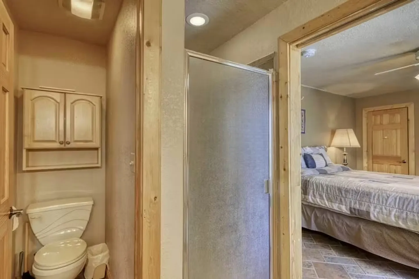 Bathroom with a toilet, wall cabinet, and frosted glass shower stall next to a doorway into an adjacent bedroom.