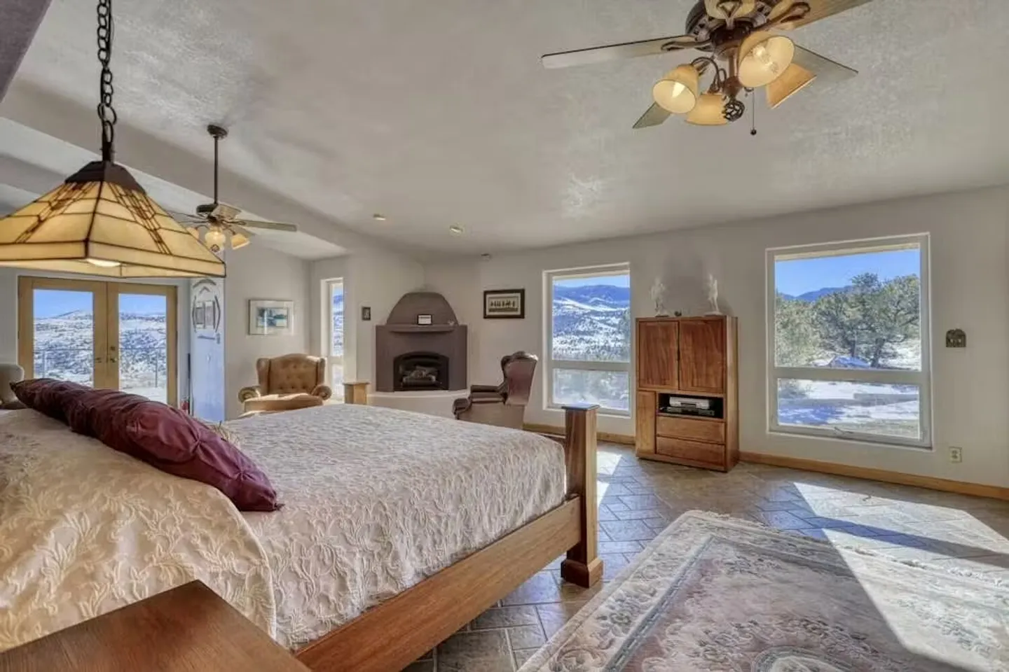 Spacious bedroom with a wooden bed, tile floors, corner fireplace, and large windows looking out to snowy hills.