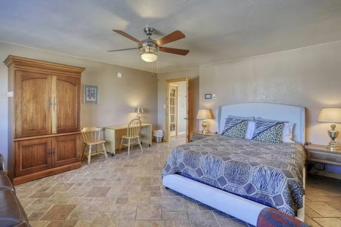 Bedroom featuring tiled flooring, a bed with a white headboard, a large wooden armoire, and a desk with chairs.
