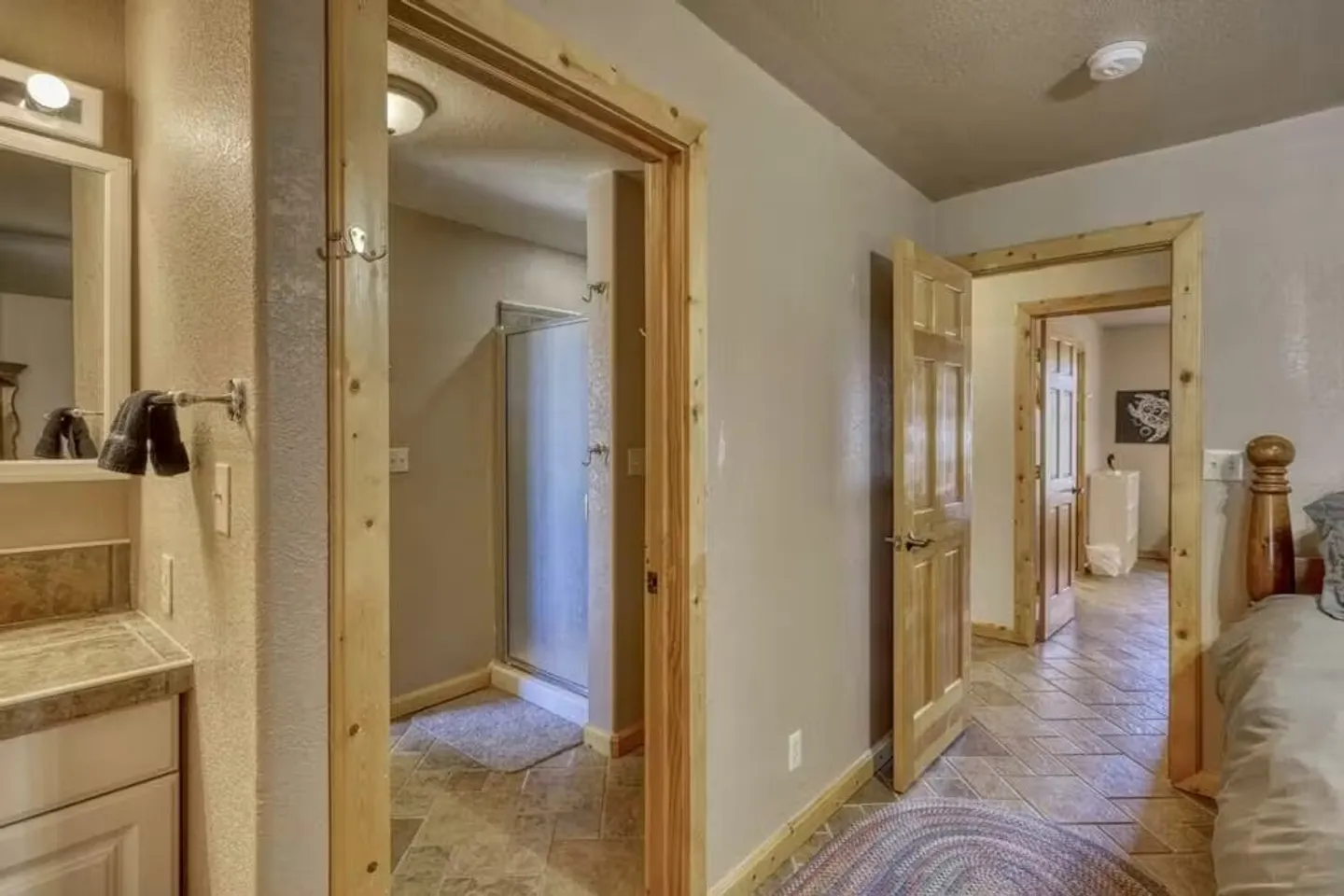 View from a bedroom looking through open doorways into an ensuite bathroom with a glass-door shower and vanity, and toward an adjacent hallway.
