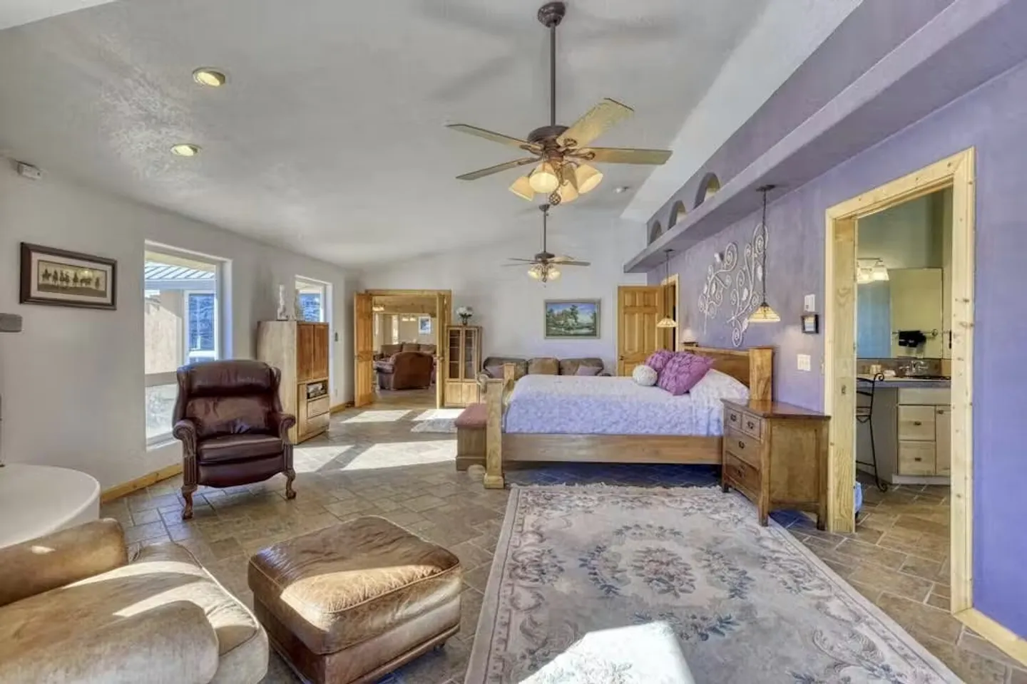 Spacious bedroom suite with tiled floors, a wooden bed frame, purple accent wall, ceiling fans, leather seating, and an adjacent bathroom.