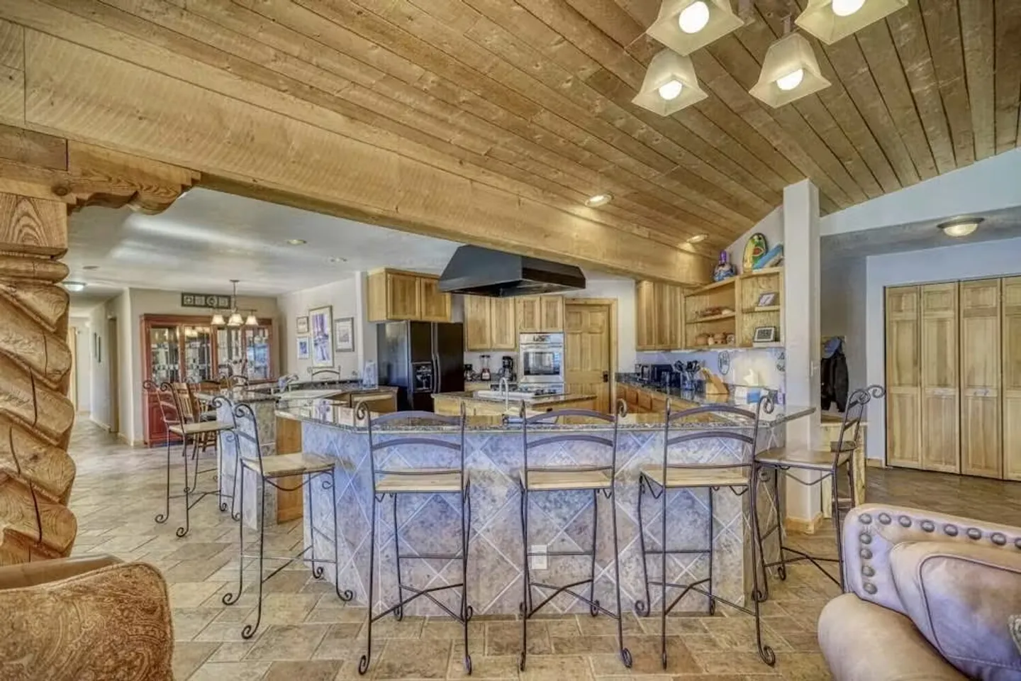 A kitchen with a curved breakfast bar, barstools, wooden ceiling, and tile flooring viewed from an adjacent living area.