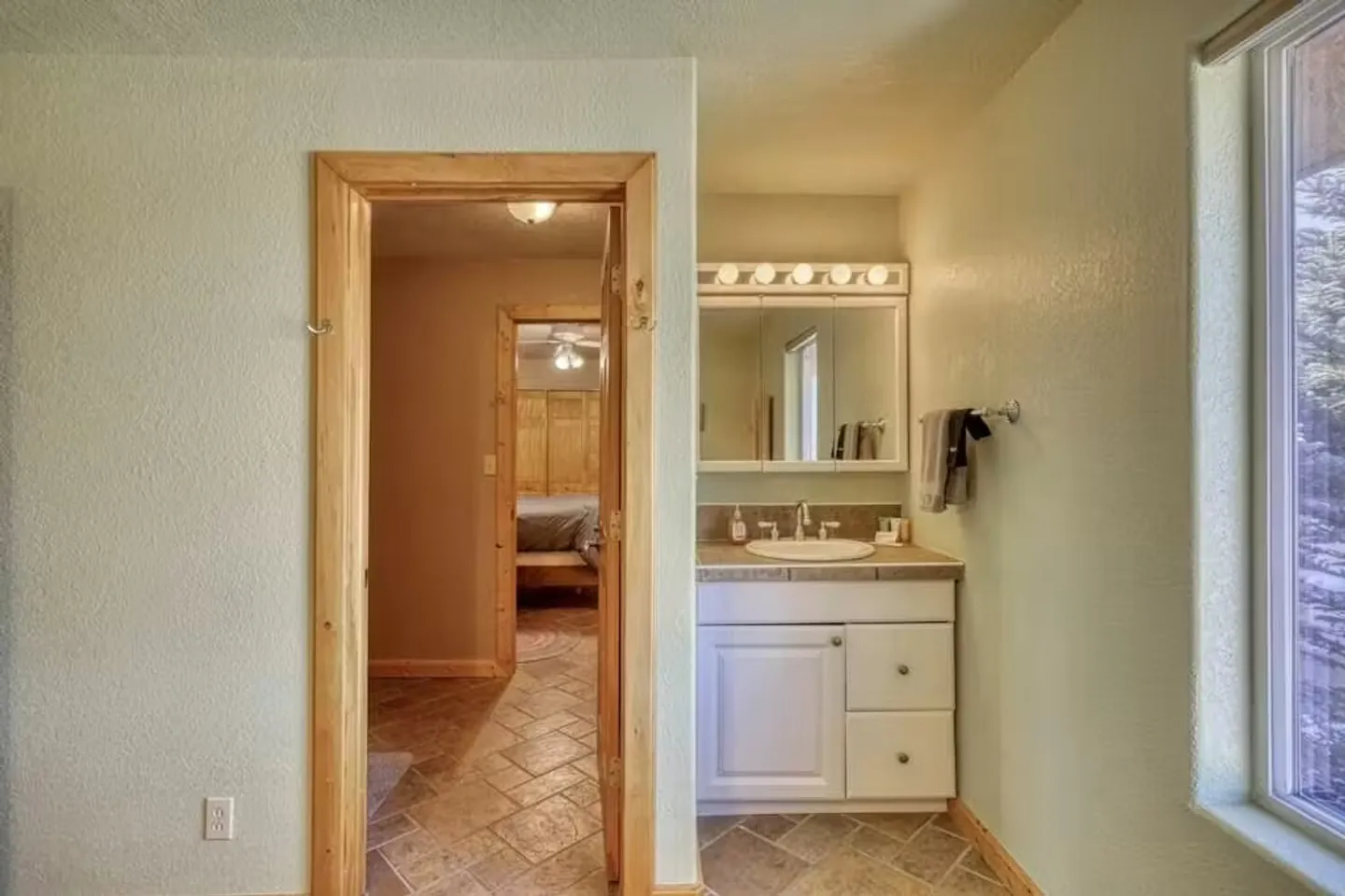 Bathroom vanity with a tiled countertop, white cabinets, and vanity lighting adjacent to an open doorway showing a bedroom beyond.