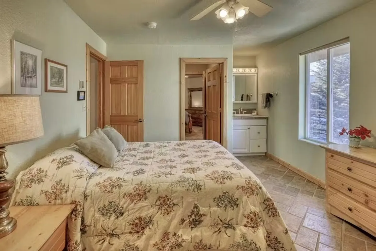 Bedroom with a patterned floral quilt on the bed, tiled flooring, wooden doors, a ceiling fan, and a view into an attached bathroom vanity area.