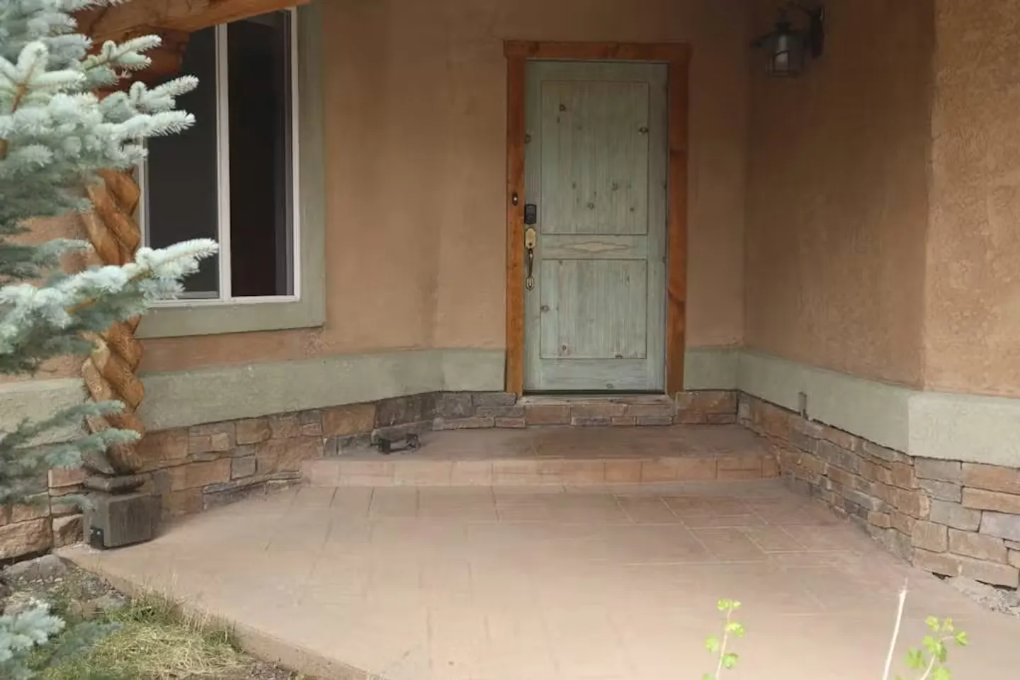 Front entrance porch of a home featuring a light green wooden door, stucco walls, stone wainscoting, and a carved wooden support column.