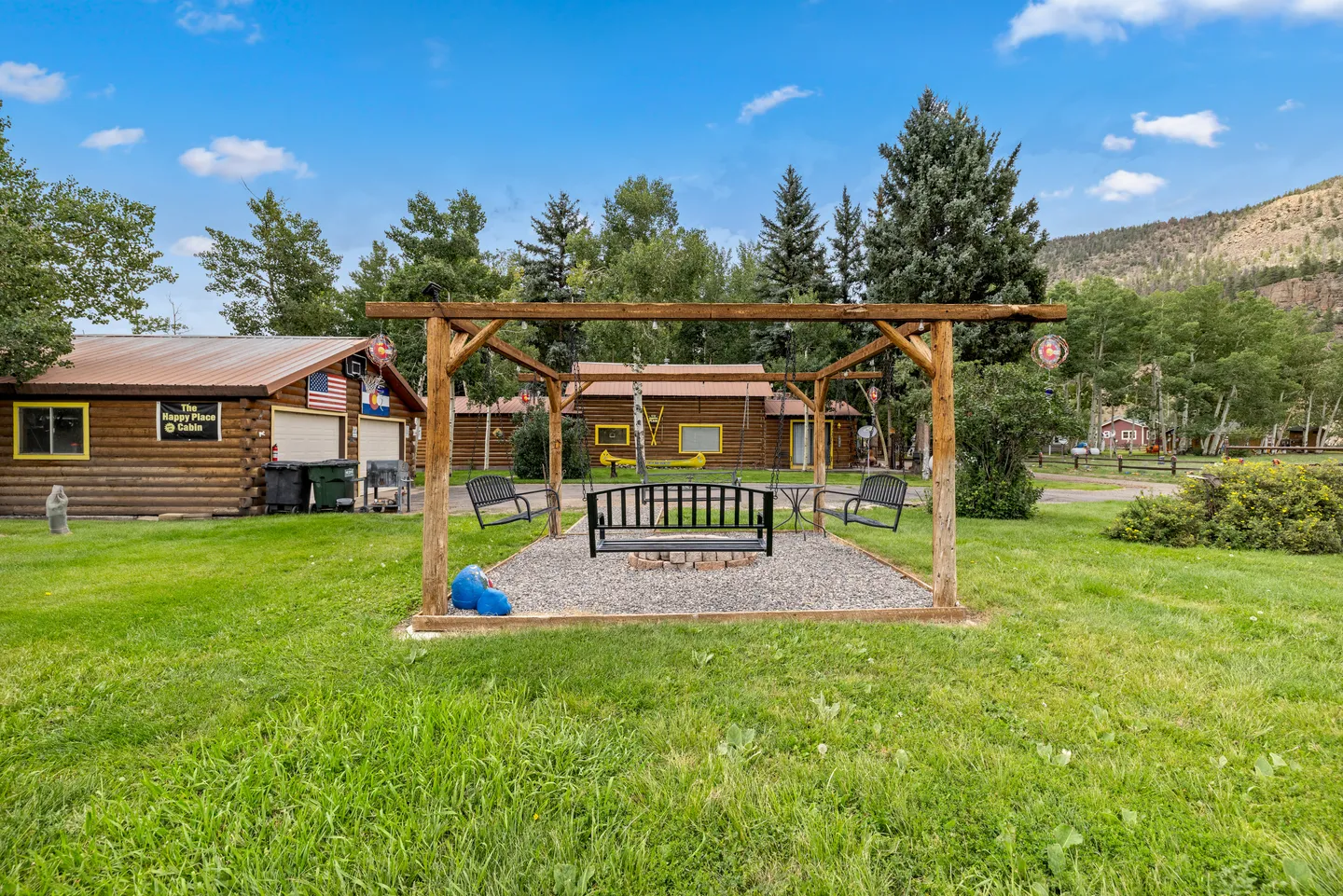 Outdoor wooden pergola framing a gravel fire pit area with hanging swing benches on a grassy lawn.