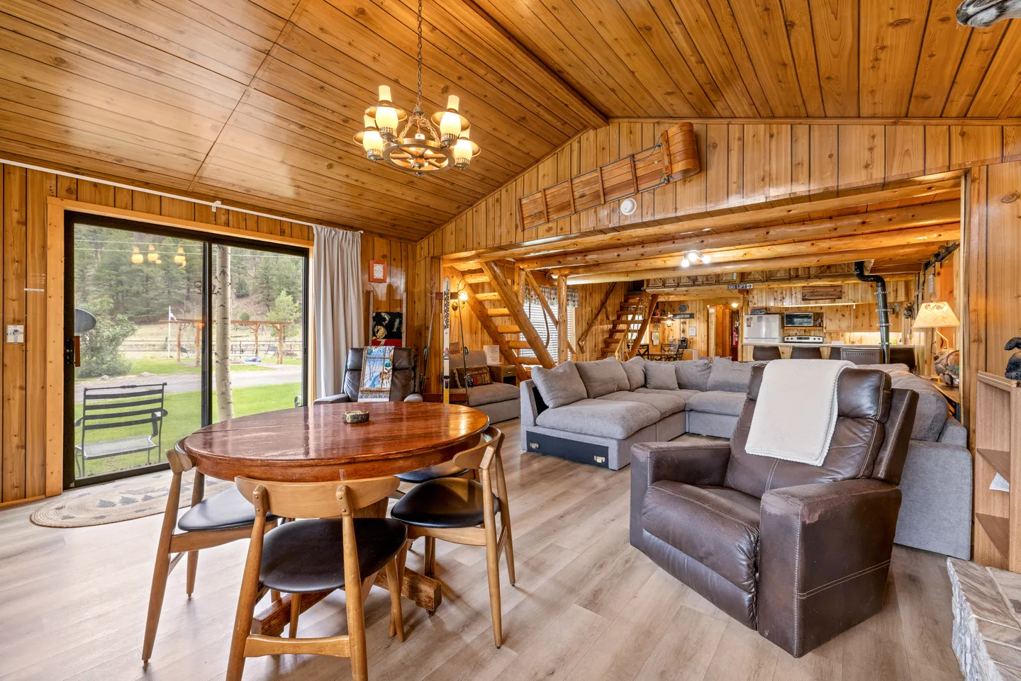Open-concept cabin interior featuring wood-paneled walls and ceiling, a round dining table, leather recliner, and large sectional sofa.
