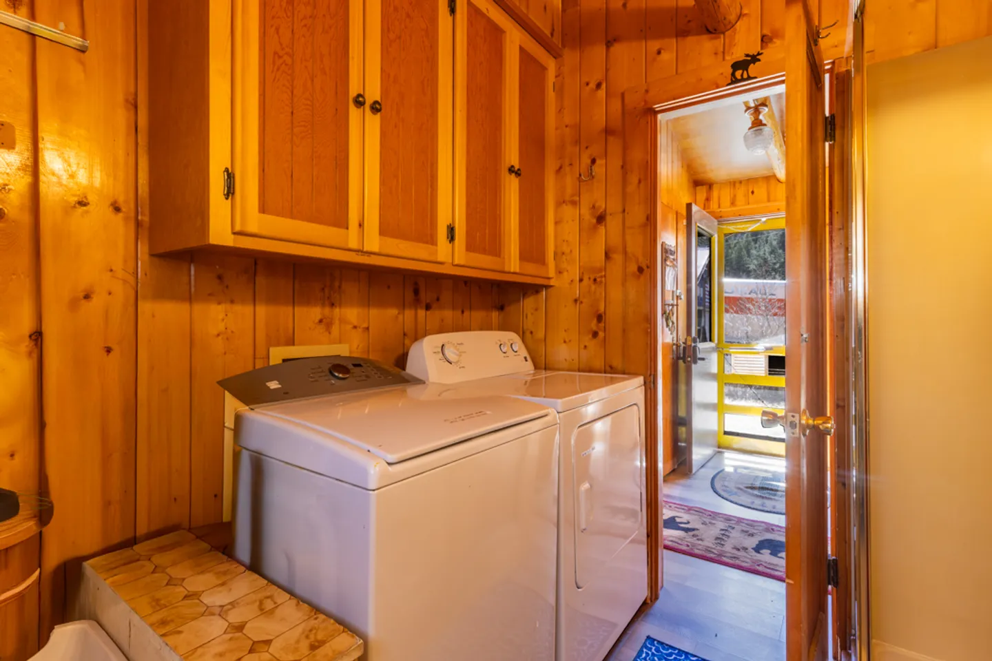 A laundry area with wood-paneled walls, upper cabinets, and a white washer and dryer next to a doorway.