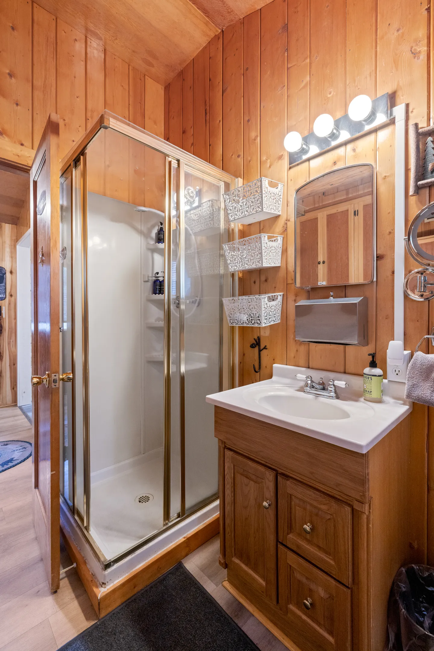 Bathroom with wood-paneled walls, a glass-door corner shower stall, and a wooden vanity with a white sink and lighted mirror.