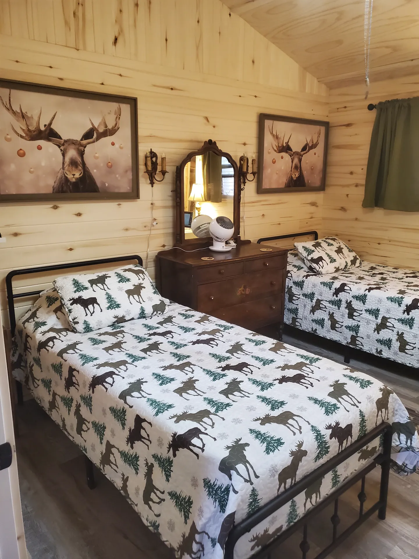 Bedroom with knotty pine wood walls, two twin beds with moose and pine tree bedspreads, and a wooden dresser with a mirror.