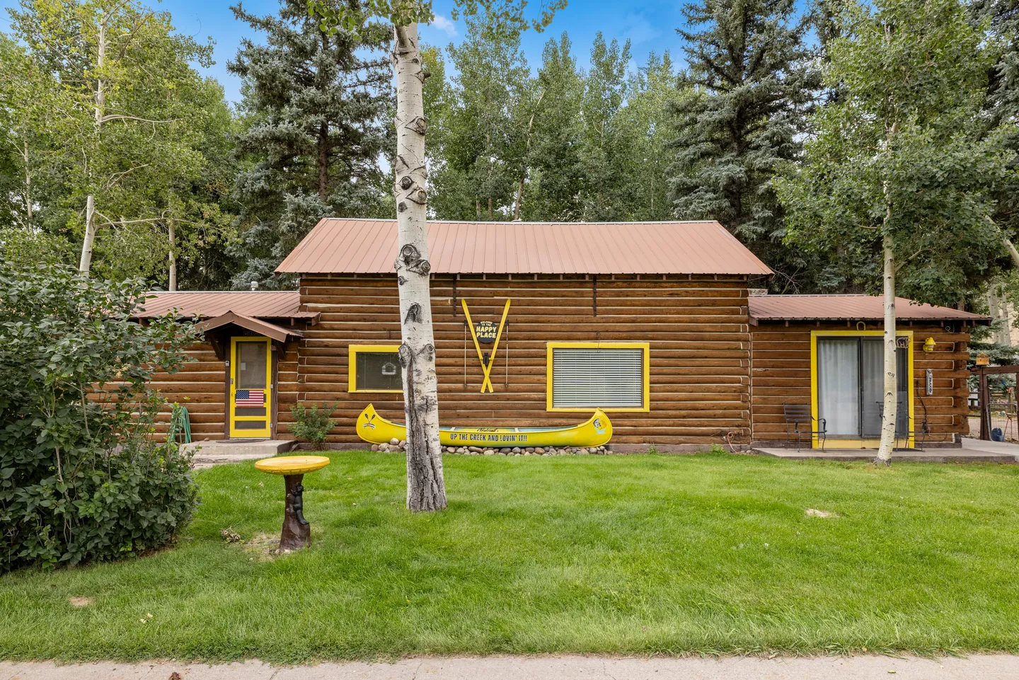 Exterior of a rustic log cabin with bright yellow trim, a yellow canoe decoration on the lawn, and a grassy yard backed by tall trees.