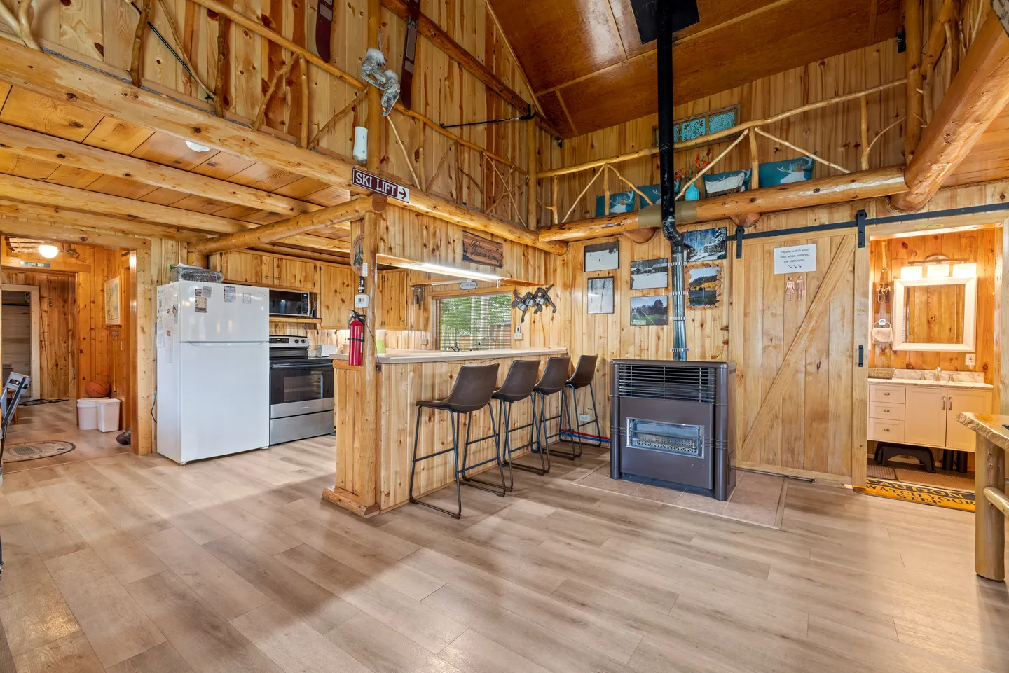 Open-concept cabin interior featuring natural wood plank walls, kitchen counter with bar stools, wood stove, and a view into a bathroom.
