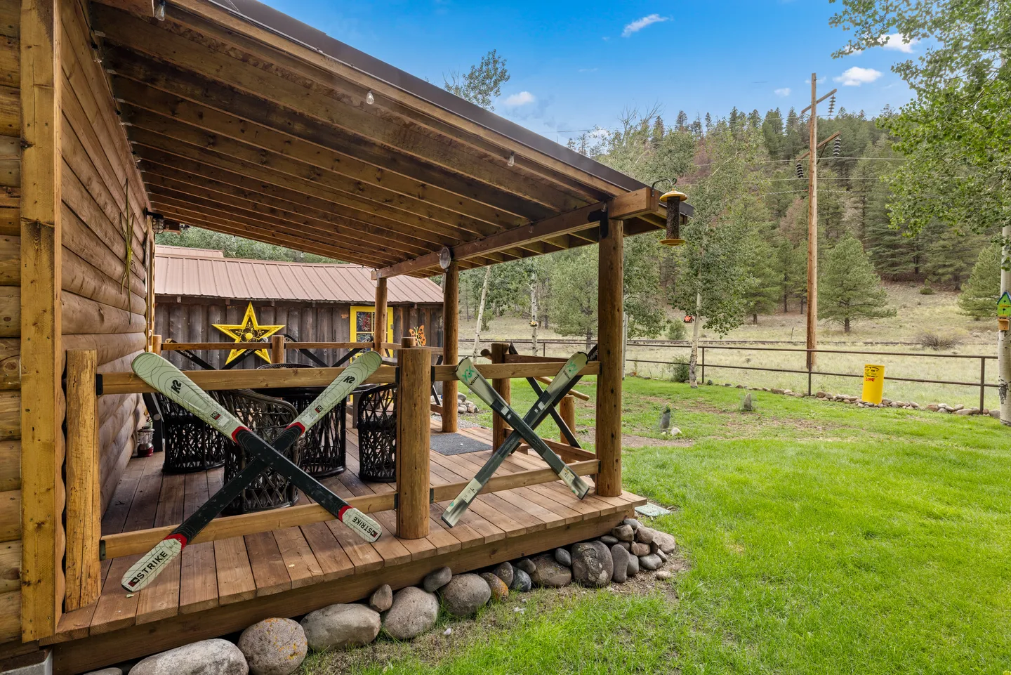 A covered wooden porch of a log cabin with crossed skis integrated into the railings, overlooking a grassy yard and forested hillside.