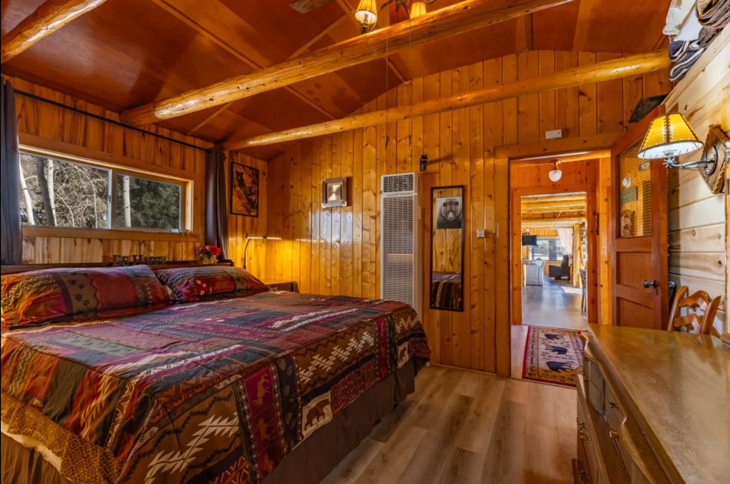 Bedroom with knotty pine wood-paneled walls, exposed log rafters, a bed with a patterned quilt, and an open doorway leading to another room.