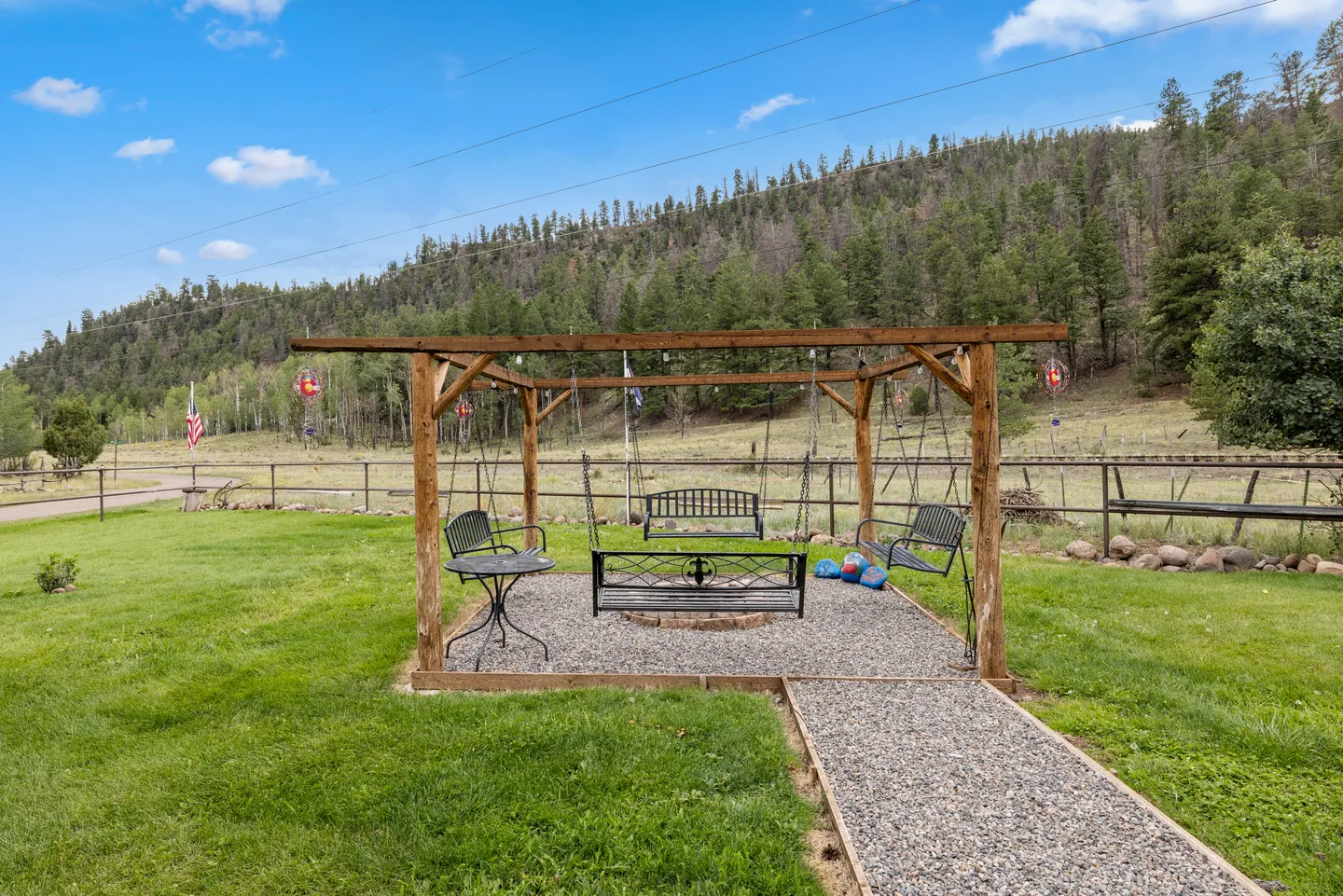 Outdoor gravel patio featuring a wooden pergola frame with hanging swing benches surrounding a central pit, set against a grassy yard and forested hillside.