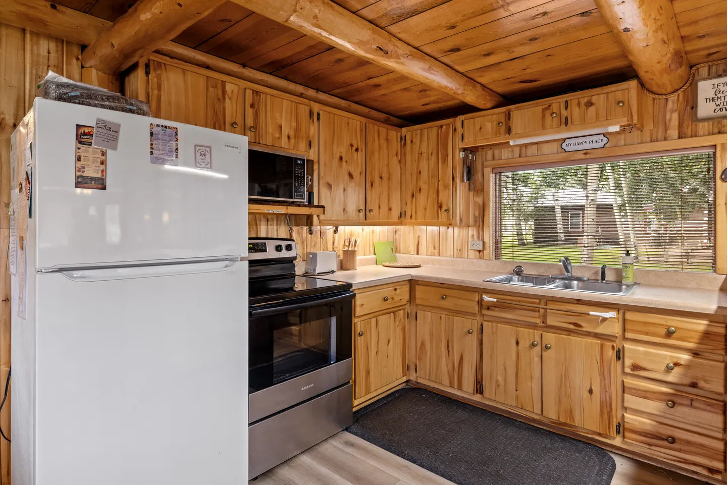 Cabin kitchen featuring knotty pine wood cabinetry, exposed log ceiling beams, a double sink under a window, and a white refrigerator.