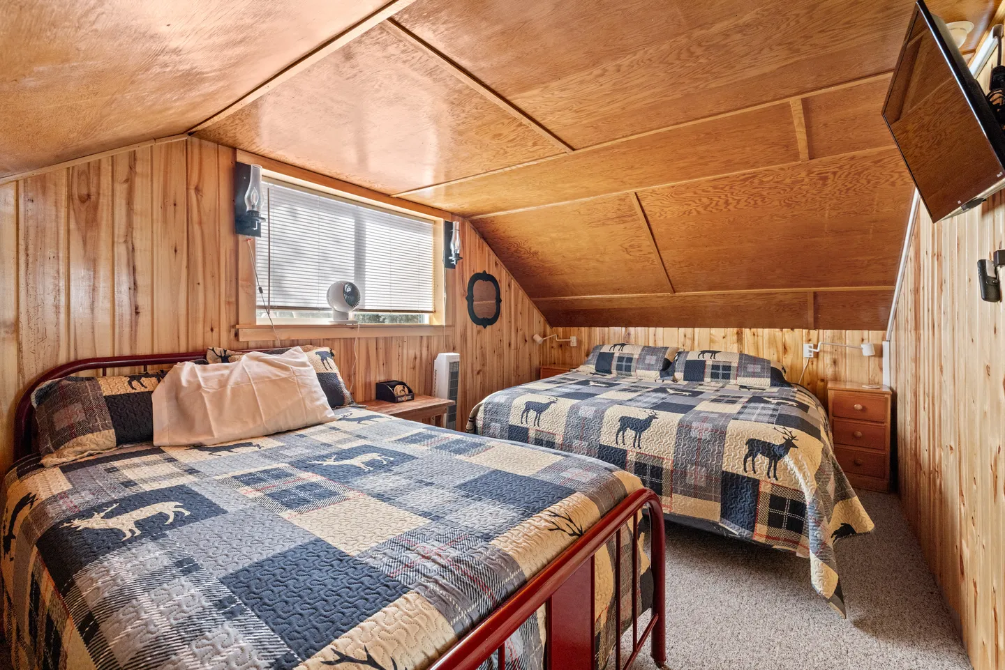 Rustic bedroom with wood-paneled walls and ceiling featuring two beds with patchwork deer quilts and a window with blinds.