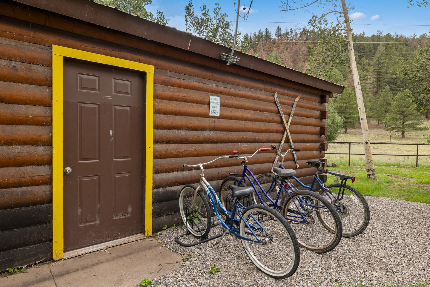 Exterior of a log-sided outbuilding with a brown door trimmed in yellow and bicycles parked in a bike rack along the gravel.