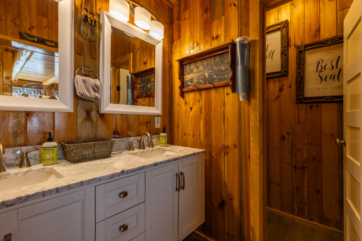 Bathroom with wood-paneled walls, a double-sink white vanity with marble-pattern countertop, and two framed mirrors.