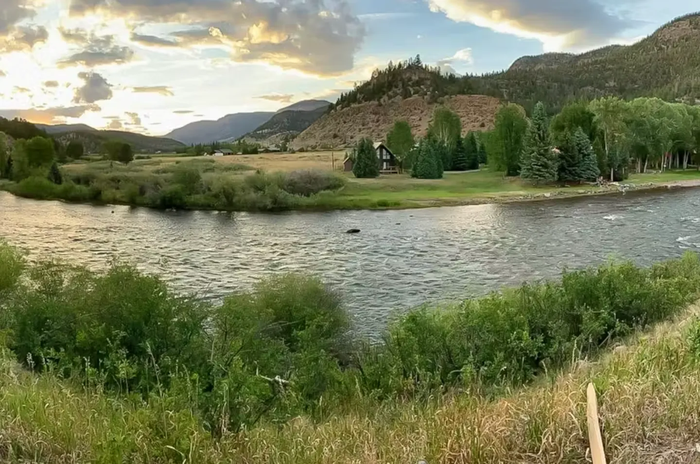 Wide scenic view of a river curving past grassy fields and distant houses beneath rolling hills and a cloudy sky.