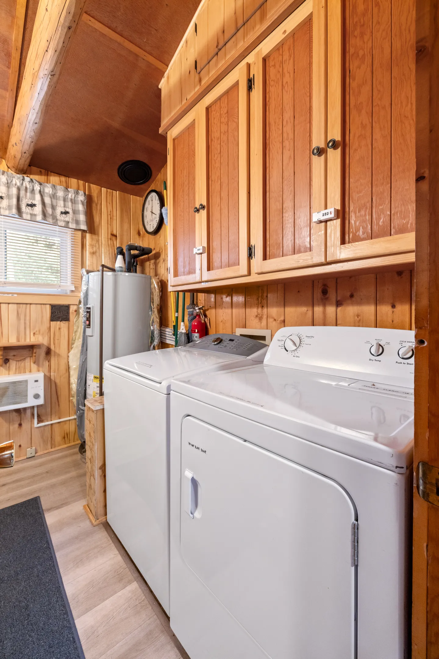 A laundry room with wood paneled walls, featuring a white washing machine, dryer, upper wooden cabinets, and a water heater in the corner.