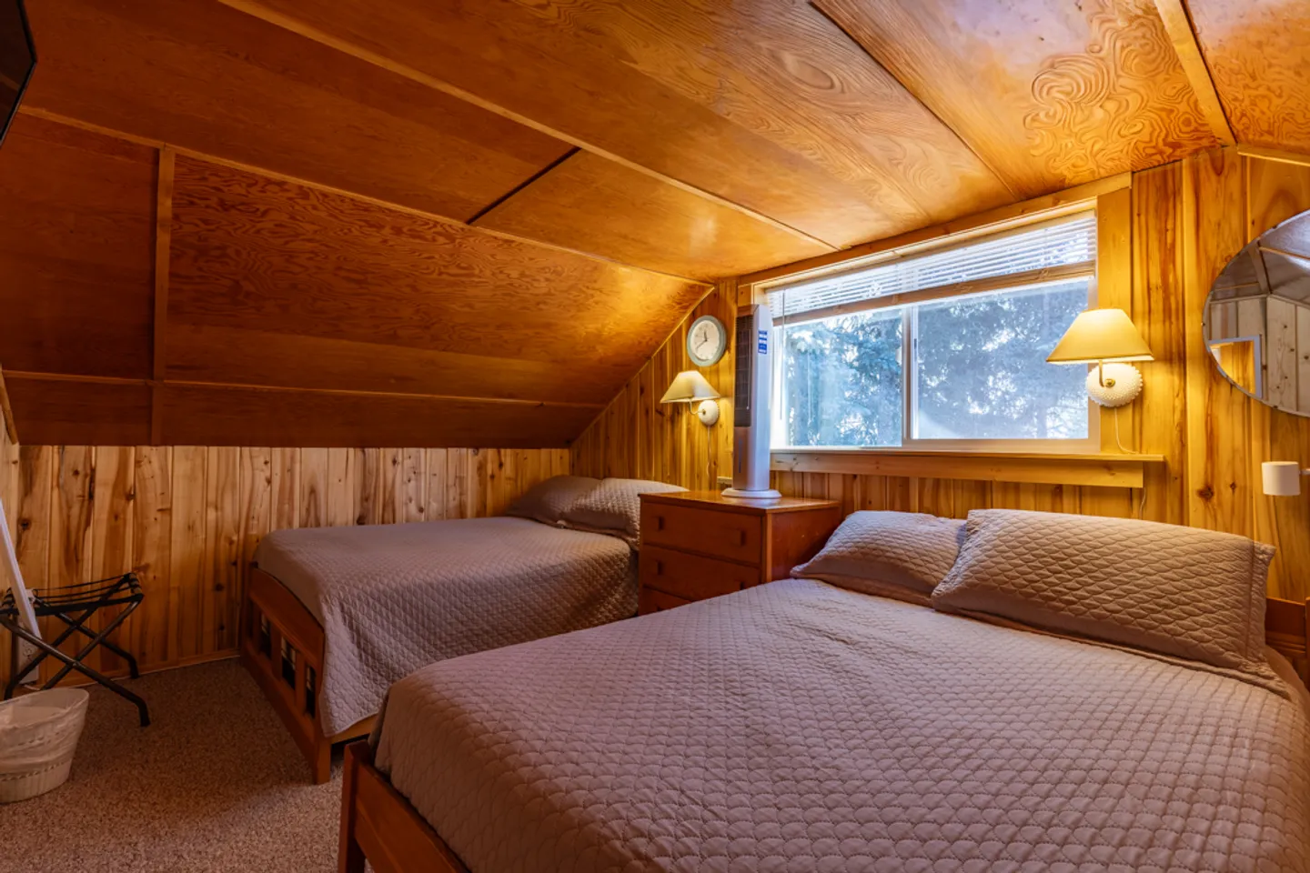 Attic bedroom with wood-paneled walls and ceiling, featuring two beds with matching quilted covers, a wooden nightstand, and a window.