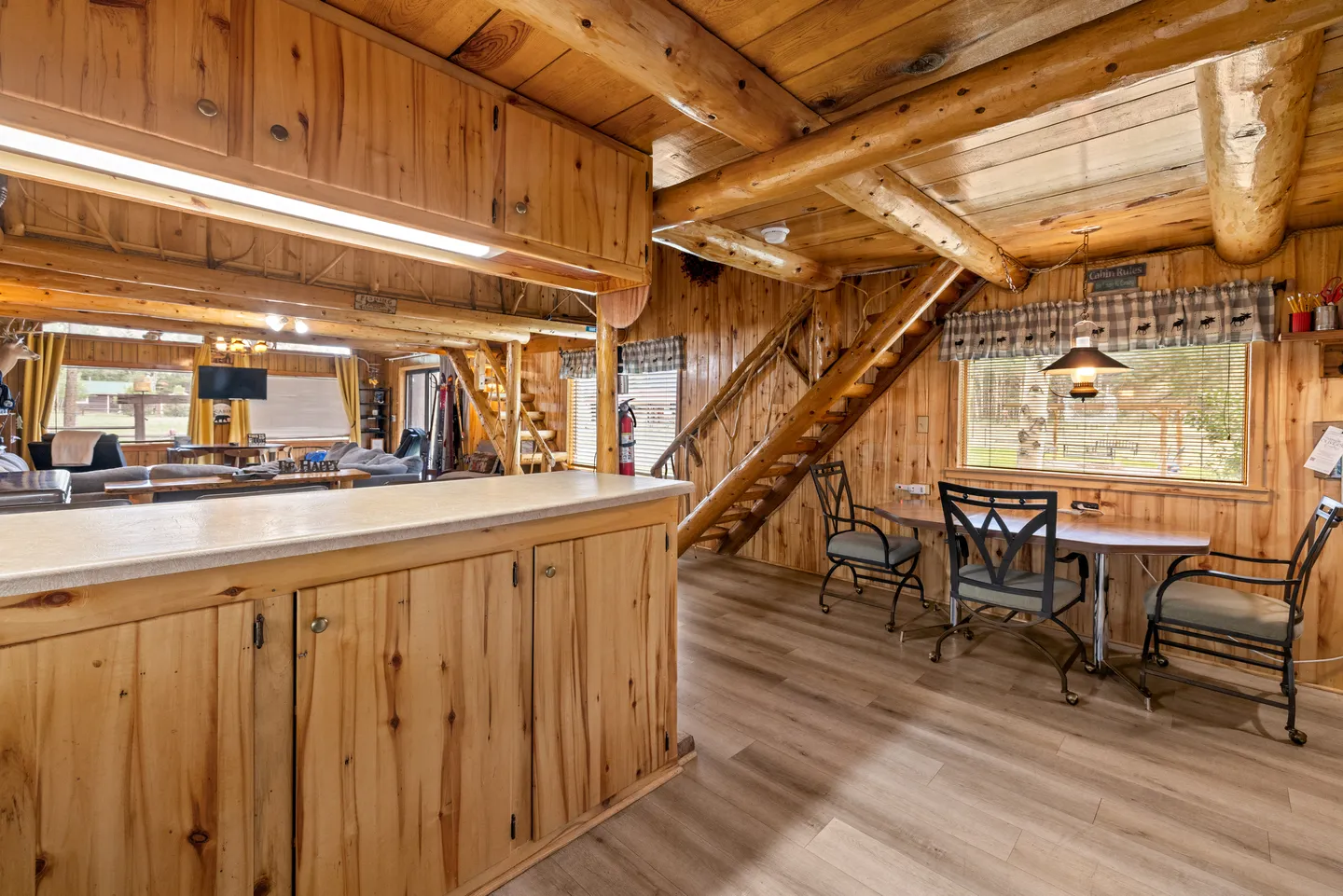 Open-concept cabin interior showing a kitchen counter, a small dining table with chairs by a window, log beams, and a staircase leading upstairs.