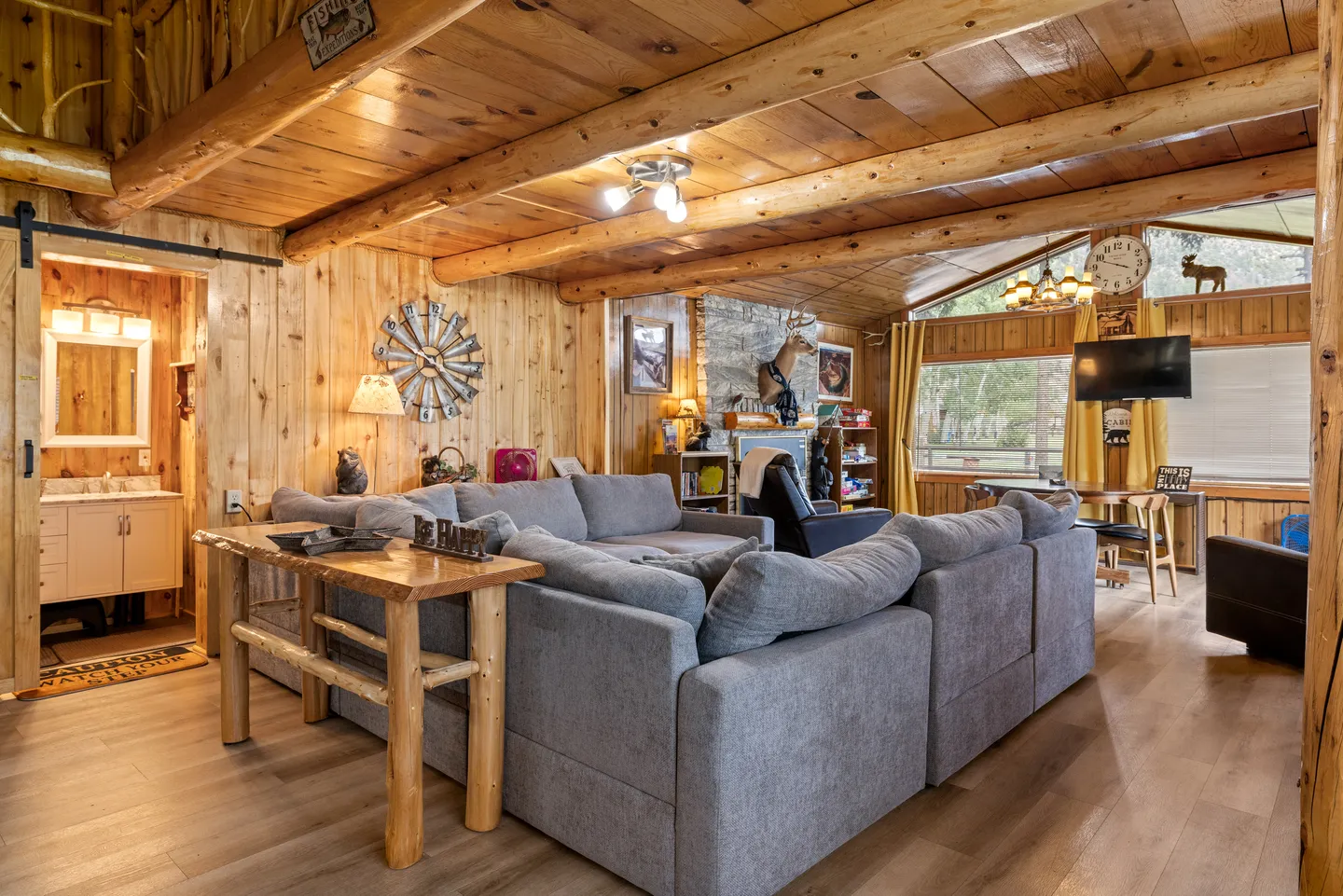 A rustic living room with knotty wood paneled walls, log ceiling beams, and a large gray sectional sofa facing an open plan area.