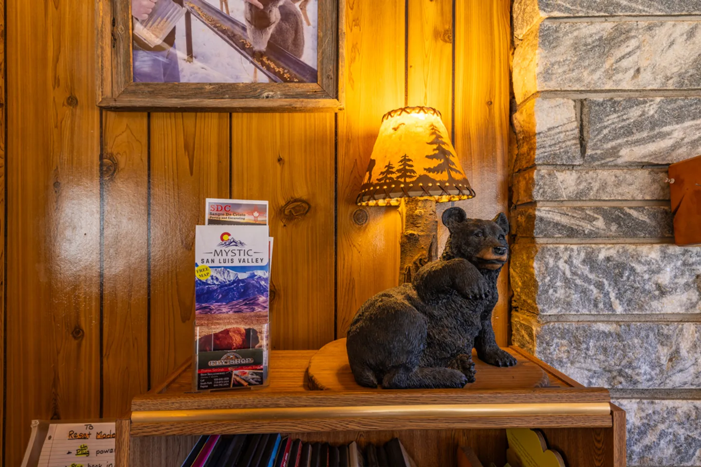 A close-up of a rustic black bear table lamp with a forest-print shade sitting on a shelf next to local travel brochures, against wood-paneled and stone walls.