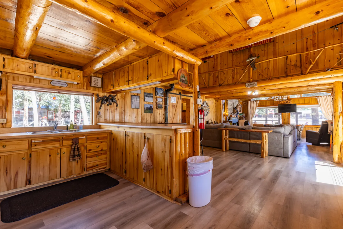 Open-concept cabin interior showing wooden kitchen cabinets, log support beams, and an adjacent living room area.