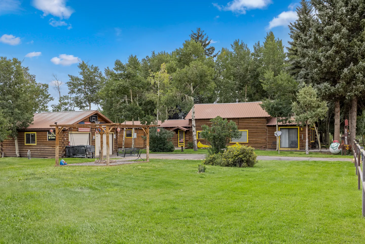 Exterior view of log-style buildings and a large green lawn bordered by tall trees under a partly cloudy sky.