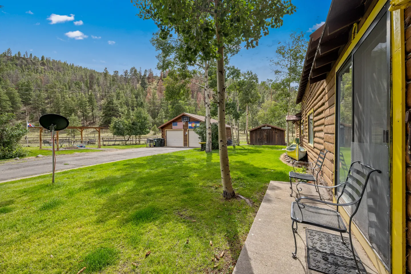 Outdoor patio along a log cabin exterior looking across a green lawn toward detached outbuildings and a forested hillside under a blue sky.
