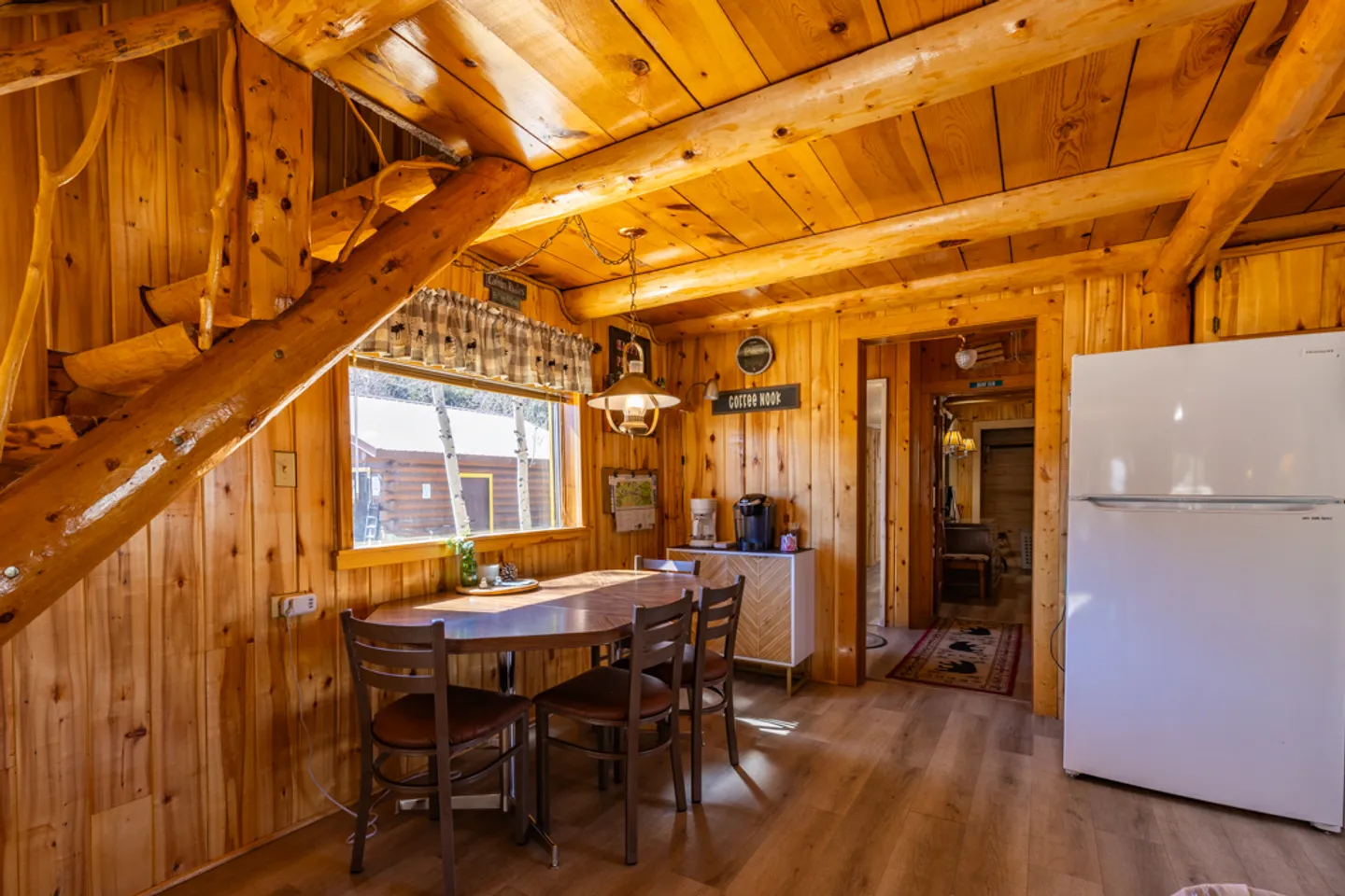 Dining area with wood paneled walls, log ceiling beams, rustic stairs, a dining table with chairs, and a white refrigerator.