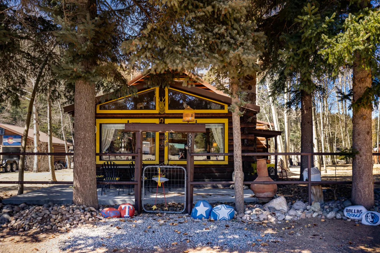 Exterior view of a rustic cabin with yellow window trim framed by tall evergreen trees behind a wooden fence with a small gate.