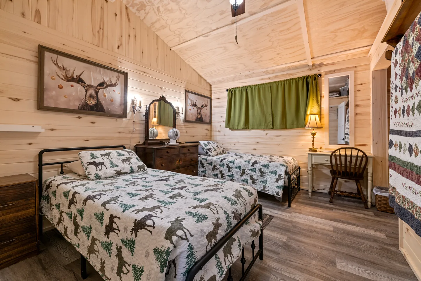 Bedroom with light wood paneled walls, two twin beds with matching moose print quilts, a vanity desk, and moose artwork on the wall.