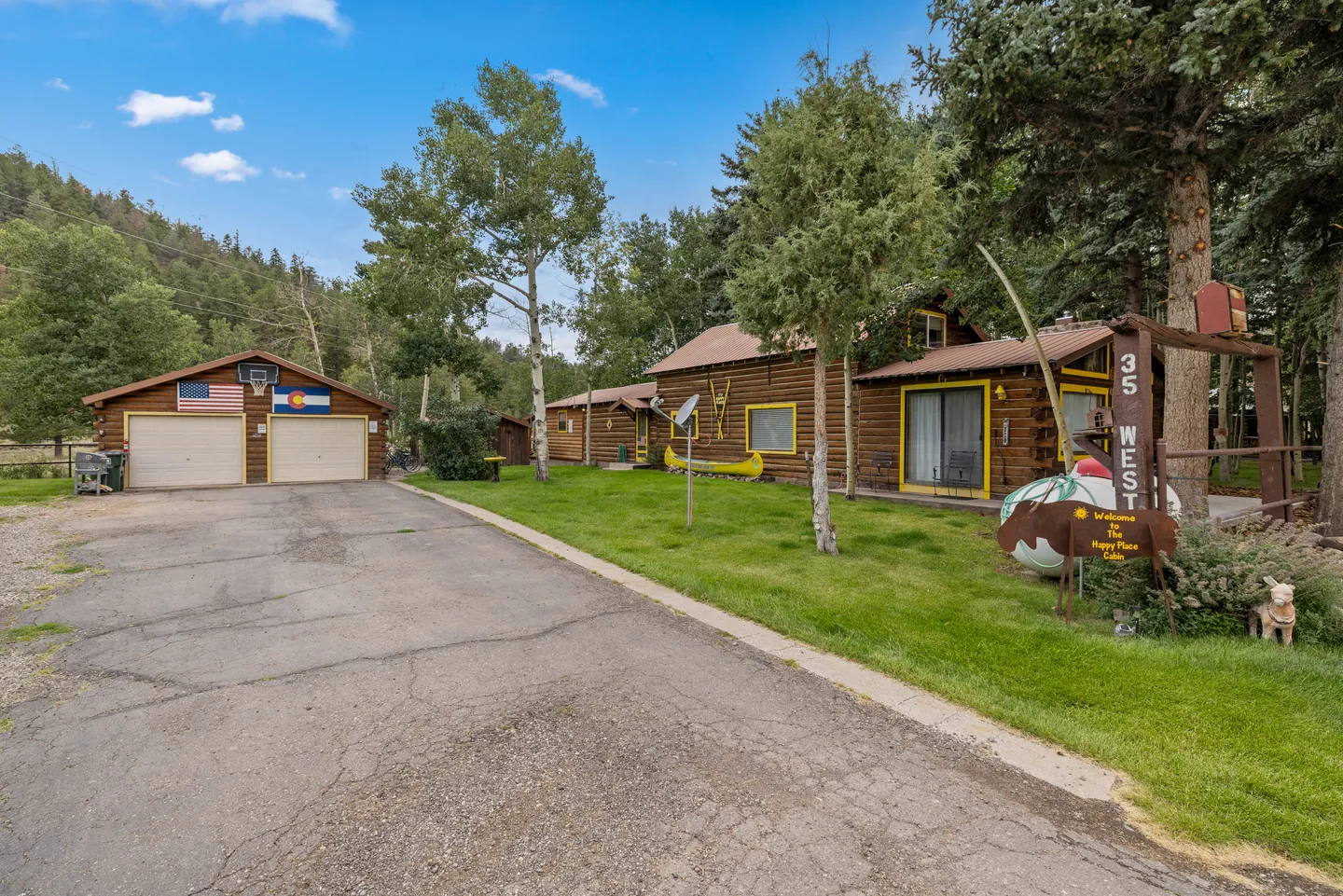 Exterior view of a log cabin with a paved driveway, detached two-car garage, and surrounding evergreen and aspen trees.