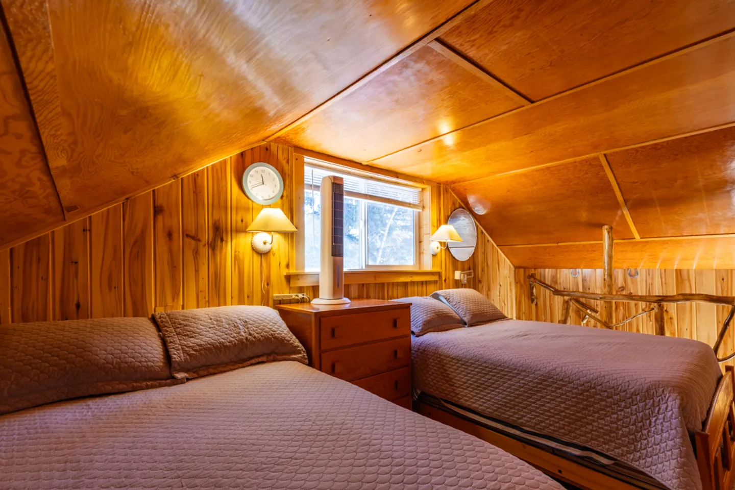 Loft bedroom with sloped wood-paneled ceilings, two beds, a small dresser, and rustic wood railings.