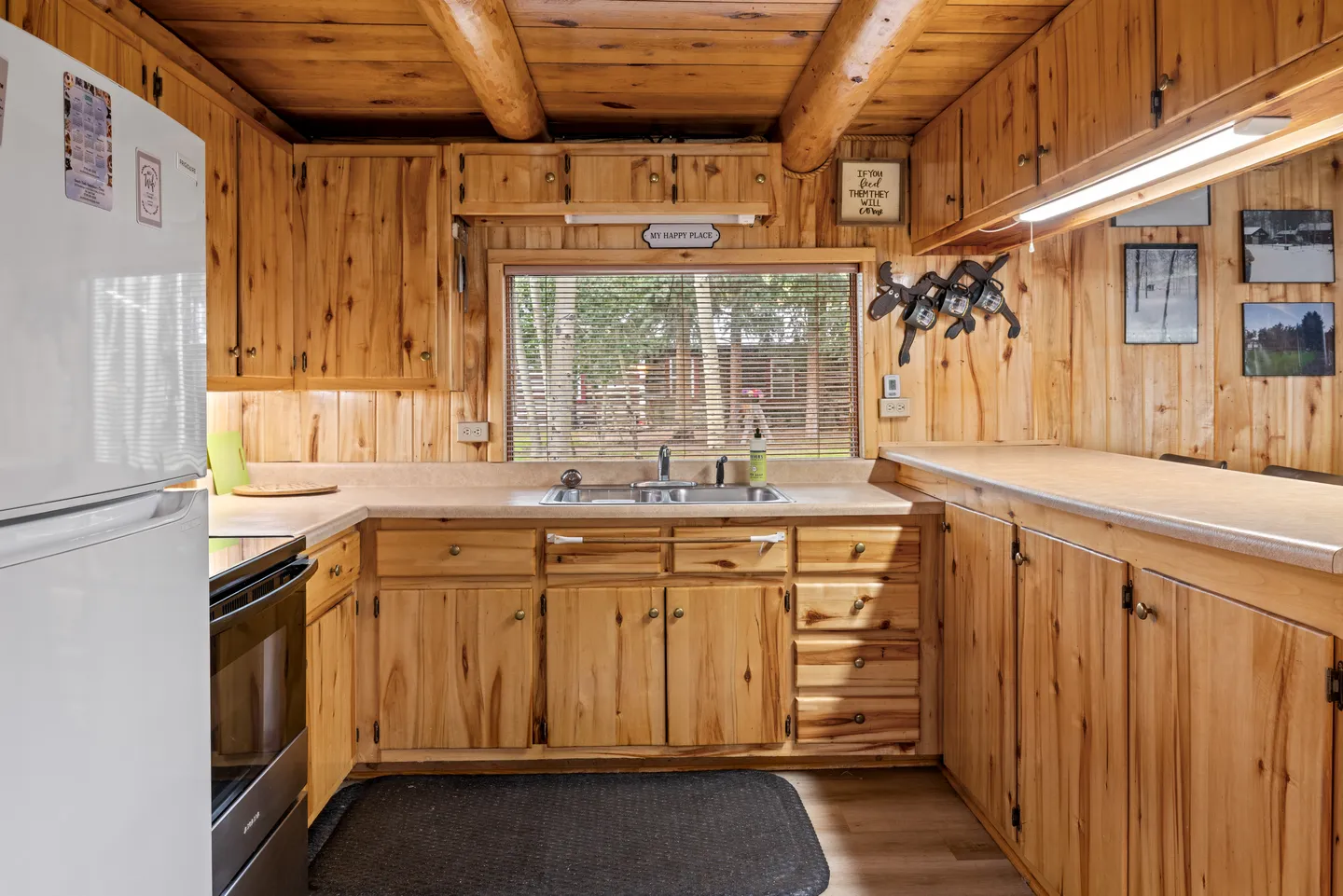 Kitchen interior featuring wood panel walls, natural wood cabinets, a laminate countertop with a stainless steel sink, and exposed log ceiling beams.