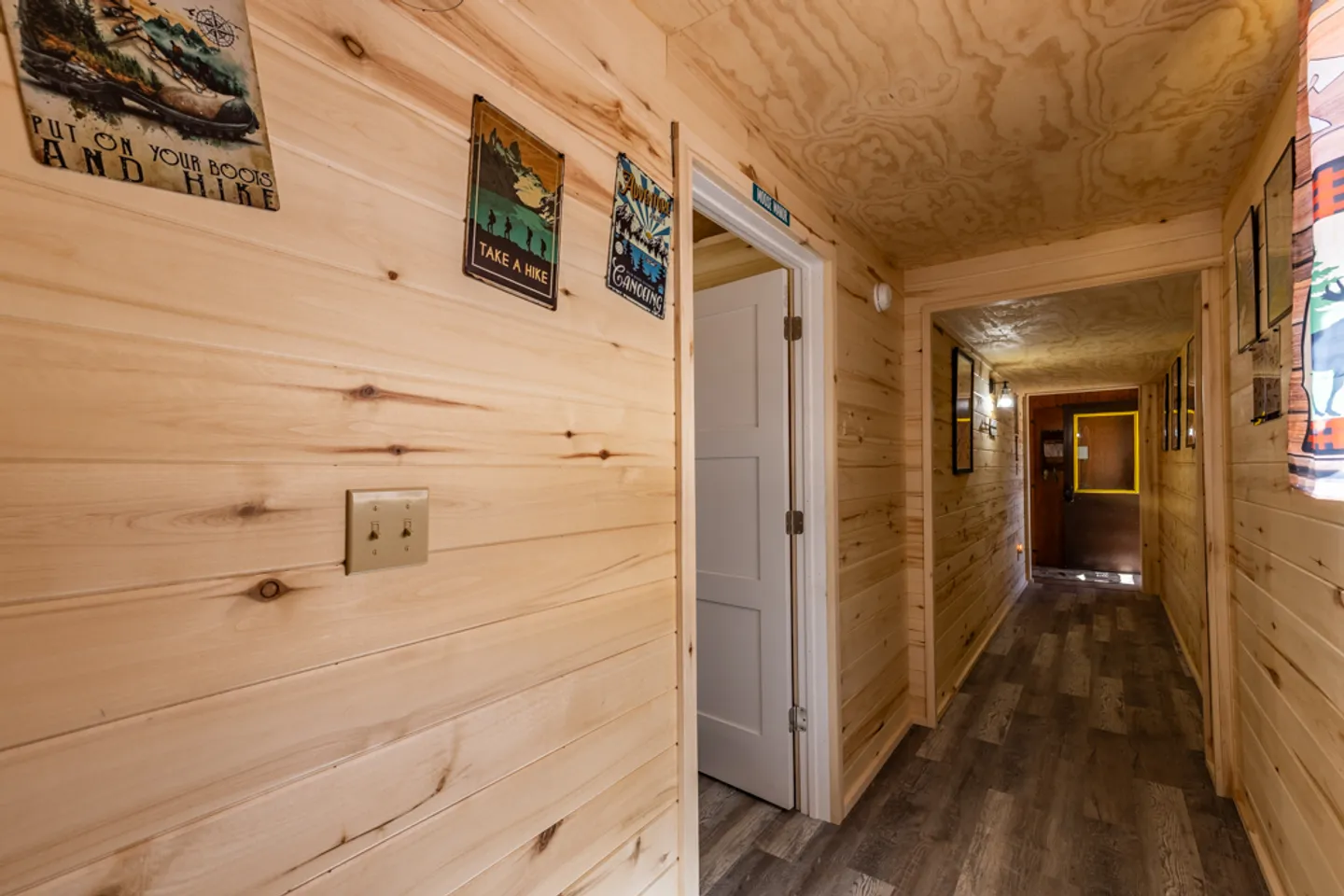 Hallway lined with light pine wood paneling, featuring wood-look floors, a white door, and framed outdoor signs.