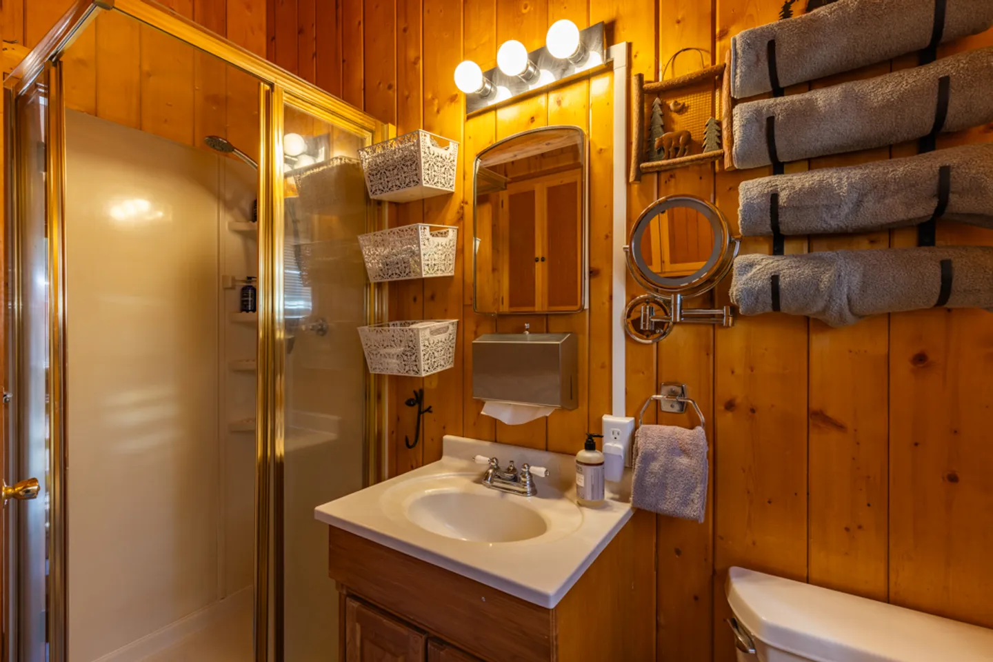 Bathroom with wood-paneled walls, vanity sink, mirror, glass-door shower, and towel rack with rolled towels.