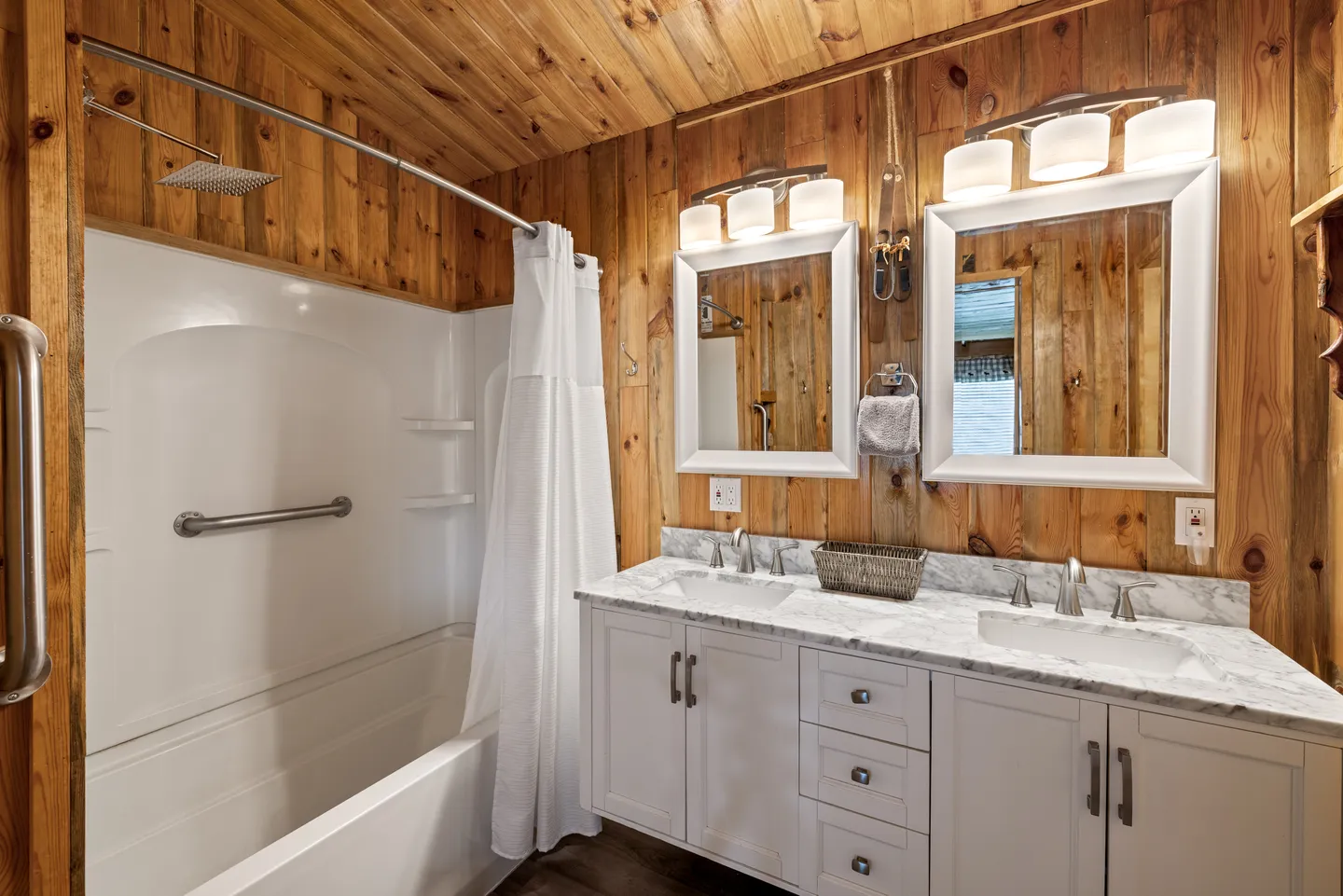 Bathroom with wood-paneled walls and ceiling, a double-sink white vanity with marble-patterned countertop, two framed mirrors, and a white shower-tub combination.