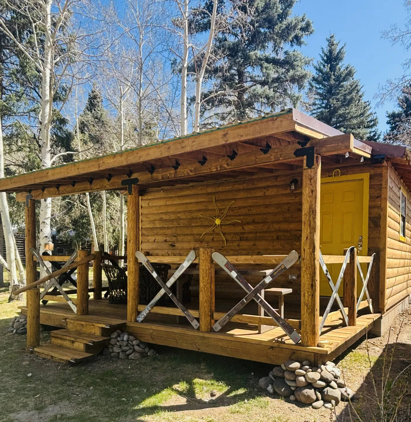 Exterior view of a small log cabin featuring a covered front porch with ski-adorned railings and a bright yellow door.