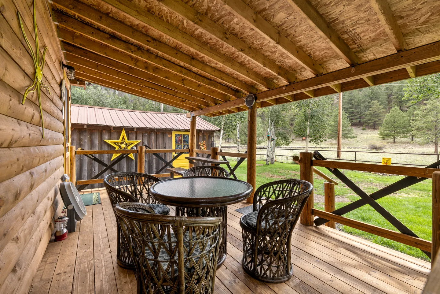 Covered wooden porch with a round outdoor table, wicker chairs, and log siding overlooking a grassy yard and outbuilding.