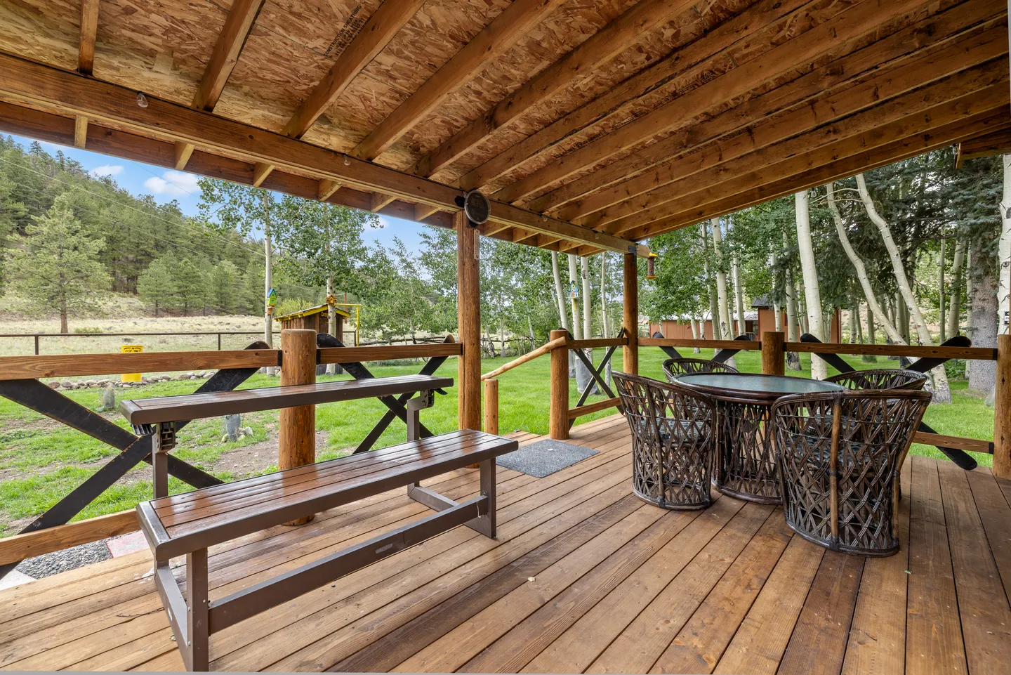 A covered wooden deck furnished with a picnic-style table and a round glass-top table with wicker chairs, overlooking a grassy yard with trees.